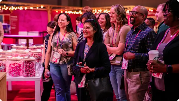 A group of diverse attendees smiling and engaging, standing in front of a table displaying gifts or treats. The background features festive lighting and a vibrant pink color scheme, creating a cheerful atmosphere at an event or conference.