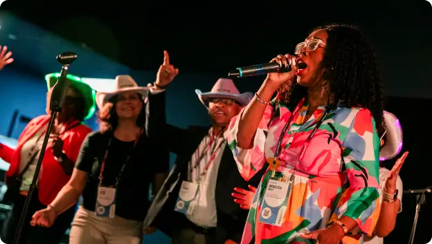 A woman singing passionately into a microphone, surrounded by a group of people in the background who are also engaged and lively. Some members of the group are wearing hats, and everyone has badges visible. The setting appears to be a festive event, with colorful lighting enhancing the vibrant atmosphere.