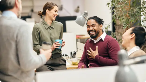 A group of four colleagues are engaged in a lively conversation in an office setting. One woman, wearing a green shirt, is smiling while holding a blue mug. A man with a beard and a burgundy sweater is laughing and placing his hand on his chest as he talks. Two other colleagues, one in the foreground holding a smartphone and another woman beside him who is also smiling, are enjoying the moment. The background features modern office elements such as desks and plants, contributing to a casual and friendly atmosphere.