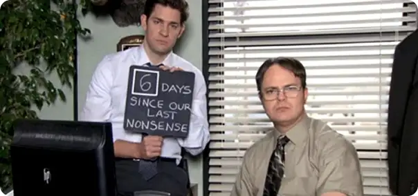 A man holding a sign that reads "6 DAYS SINCE OUR LAST NONSENSE" stands next to another man in an office setting. The first man is smiling with slight amusement, while the second man has a serious expression. The background features blinds and greenery, creating a typical office atmosphere.