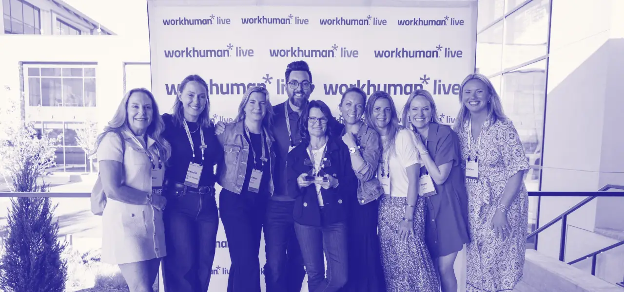 A group of eight people poses for a photo in front of a backdrop displaying "workhuman live." They are smiling and appear engaged, with one person holding an award. The setting suggests a celebratory atmosphere, likely related to a conference or event.