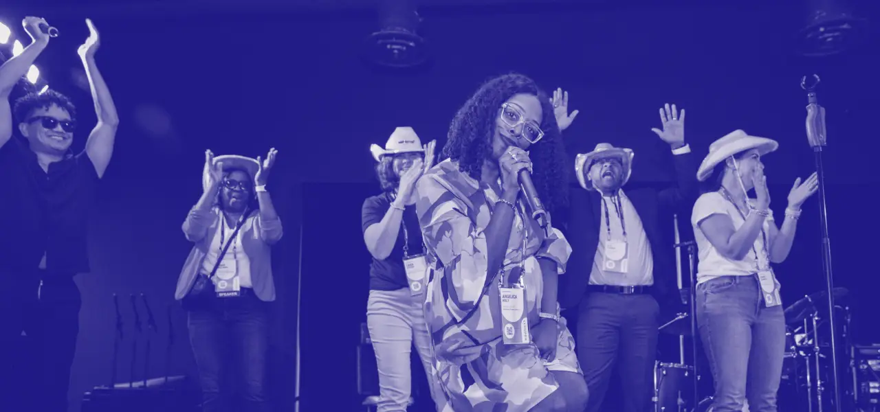 A lively scene featuring a diverse group of people on stage, enjoying a performance. A woman is at the forefront, holding a microphone and smiling while wearing a patterned outfit and glasses. Behind her, several individuals, including a man in sunglasses and others in cowboy hats, are participating enthusiastically, clapping and celebrating. The background suggests a lively atmosphere, capturing moments of joy and engagement at an event.