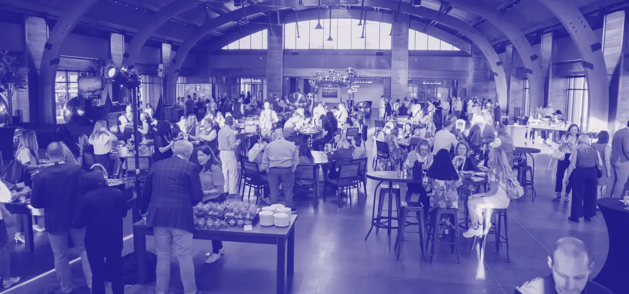 A busy event space filled with numerous attendees engaging in conversation, enjoying refreshments, and actively participating in various activities. The venue features high ceilings and large windows, providing natural light, while tables are arranged throughout the area, where guests gather. The atmosphere is lively, with decorations and elements that create a welcoming environment.