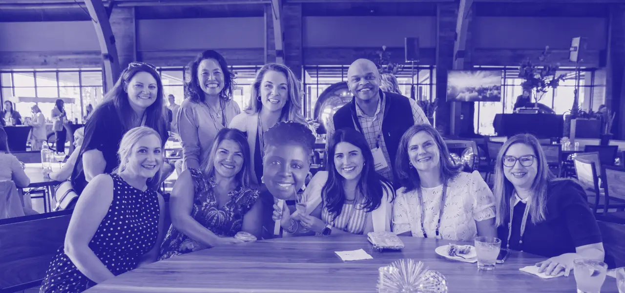 A group of nine people, including women and a man, gather around a table, smiling and posing for the camera. One woman holds a face cutout of another individual, and everyone is dressed casually. The setting appears to be a lively event in a well-lit venue, with other attendees visible in the background.