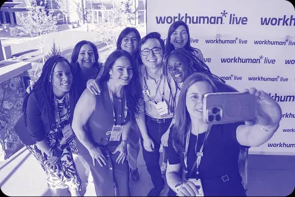 A group of eight smiling individuals take a selfie together in front of a backdrop featuring the Workhuman Live logo. They are enjoying their time at the event, dressed in casual attire. The setting appears to be outdoors with greenery visible in the background.
