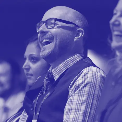 A man wearing glasses and a checkered vest is laughing joyfully. He is seated among others with bright expressions, suggesting an atmosphere of enjoyment or engagement, likely during an event or presentation. The image has a blue-toned overlay, enhancing the lively feel of the moment.