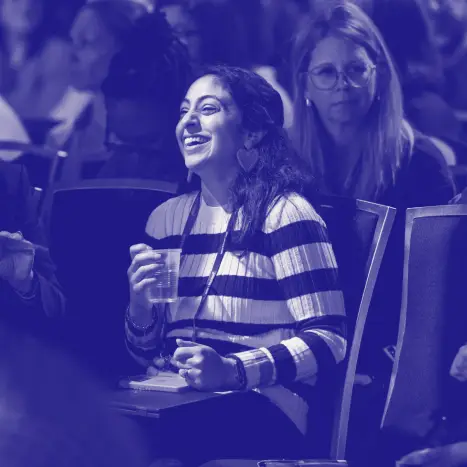 A woman with dark, wavy hair wearing a striped sweater is seated in an audience, smiling broadly while holding a cup. In the background, other attendees are visible, some appearing engaged in conversation, contributing to a lively atmosphere at an event. The scene is presented in a monochromatic blue tone.