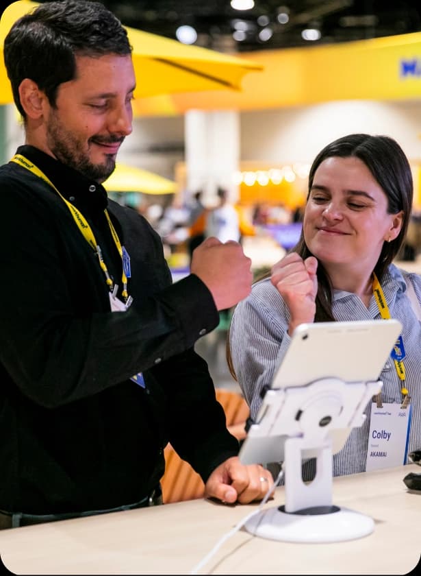 Two people, a man and a woman, are standing beside a table with a tablet on a stand. They are smiling and making fist bumps, while wearing lanyards with name tags that read "Colby AKAMAI." The background features colorful lighting and a blurred setting suggesting a busy event.