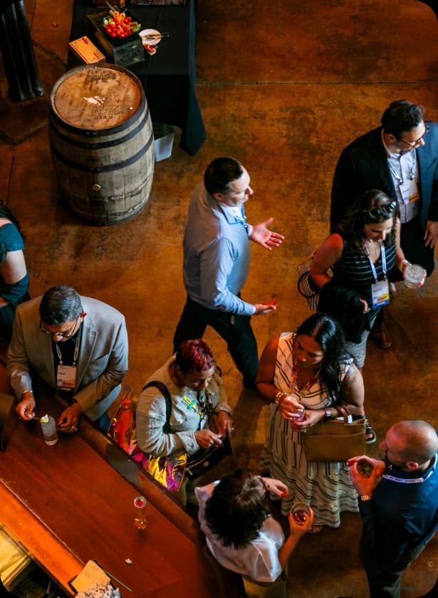 A group of people socializing at an indoor event, with some individuals holding drinks while others engage in conversation. In the background, there is a wooden barrel and a table with various items, including fruits. The floor has a rustic texture, and there are a mix of formal and casual outfits among the attendees.