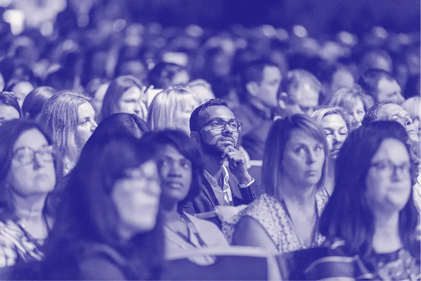 A diverse audience attentively listens during a presentation, with a focus on a man in a suit looking thoughtful among a sea of engaged faces. The image conveys an atmosphere of interest and participation, with individuals ranging from men to women, all appearing concentrated on the speaker. The overall mood is one of engagement and professionalism, set in a conference or event environment.
