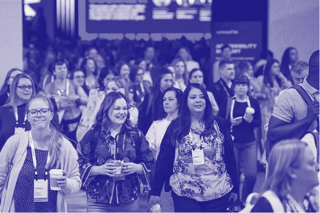 A large crowd of attendees walking through a conference venue, smiling and engaging with one another. Some individuals are holding cups, and name badges can be seen on several attendees. A digital display in the background features information related to the event, contributing to a vibrant and animated atmosphere.