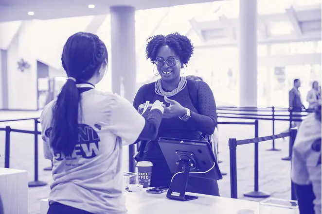 A woman smiling as she receives a wristband from a staff member at a registration desk. The staff member is wearing a shirt with the words 'Event Crew' visible. In the background, there are people waiting in line and a coffee cup labeled 'Chance Coffee.' The setting appears to be a conference or event venue.
