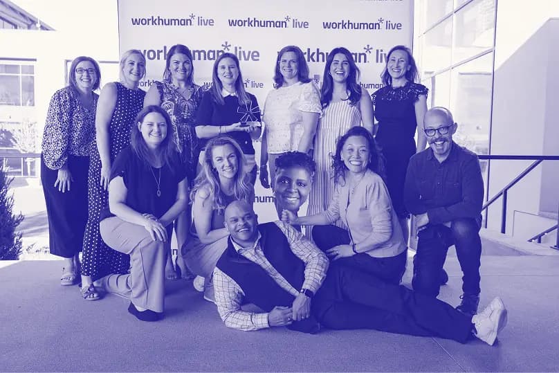 A group of diverse individuals poses for a photo in front of a backdrop that reads "workhuman live." The setting appears to be a conference or event, with some people standing and others sitting. One individual holds an award, while others display smiles, showcasing a sense of camaraderie and celebration.