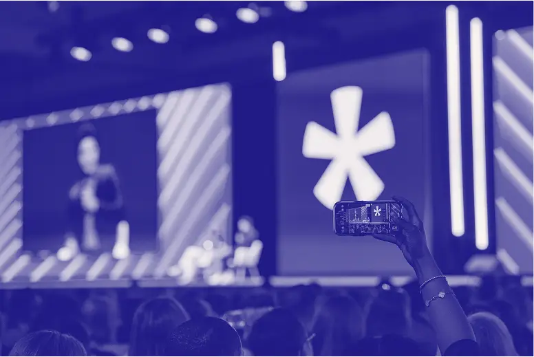 A person in the audience holding a smartphone captures a speaker on stage at a conference. The background features a large screen displaying a prominent asterisk logo, with additional lighting effects highlighting the setup. The atmosphere suggests a lively event with engaged attendees visible in the foreground.