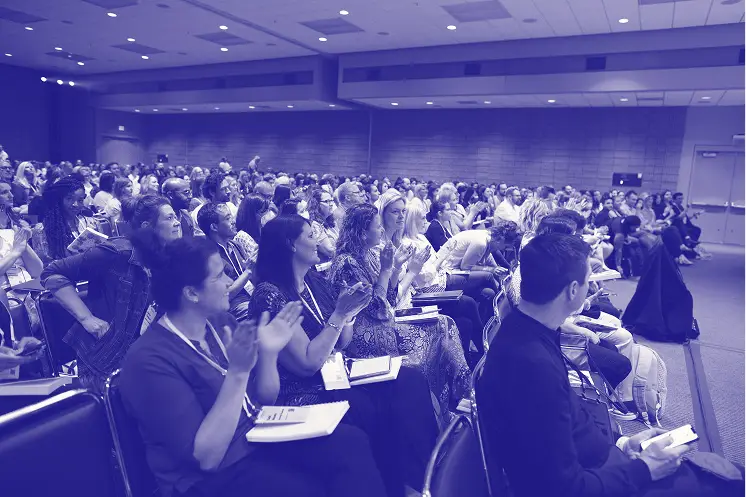 A large audience of people is seated in a conference hall. Many attendees are engaged and applauding, showing enthusiasm for the presentation or event happening on stage. The room is well-lit, with a visible stage area at the front. Notebooks and materials are on the laps of several participants, indicating they are taking notes. The atmosphere appears lively and attentive.