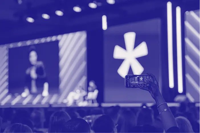 A person in the audience holds up a smartphone to capture a speaker on stage at an event. The backdrop features large screens displaying a logo, along with bright stage lights illuminating the setting. The focus is on the audience's engagement, highlighting the atmosphere of the event.