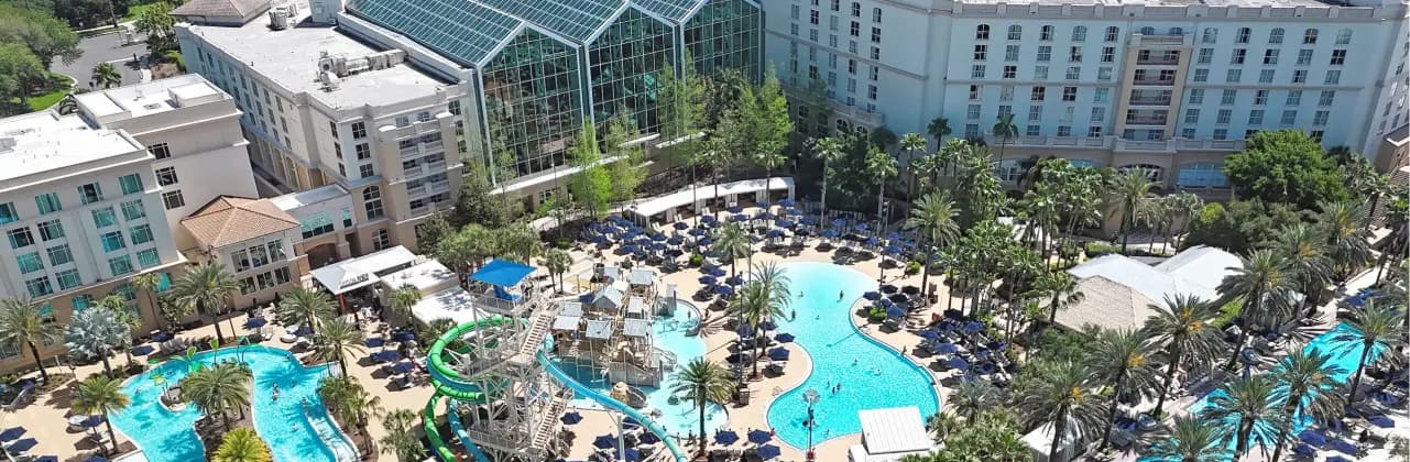 Aerial view of a resort featuring a large swimming pool area surrounded by palm trees and lounge chairs. In the background, there are several buildings, including one with a glass enclosure. The pool has a waterslide and several play structures for children, creating a lively and inviting atmosphere for guests.