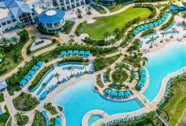 Aerial view of a resort landscape featuring winding pools and green spaces. The area includes numerous lounge chairs arranged by the water, surrounded by lush palm trees and various plantings. In the background, a large white building with a blue roof and balconies can be seen. The setting is bright and inviting, suggesting a tropical atmosphere.