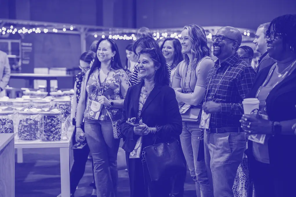 A group of attendees smiling and engaged in conversation at an event, standing in front of tables filled with jars of treats. The setting has festive decor in the background, suggesting a lively atmosphere. People are wearing lanyards, indicating their participation in the event. The image has a vibrant yet soft blue tint that enhances the joyful mood.