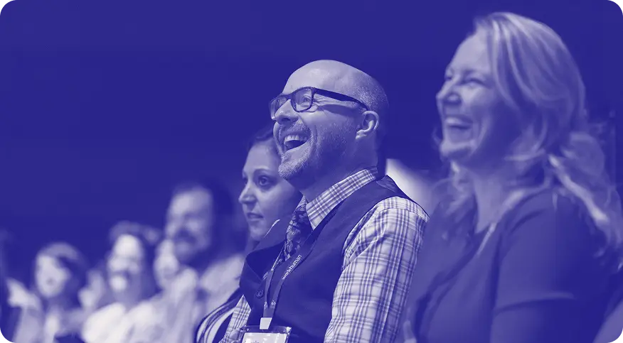 A group of people sitting together, with a focus on three individuals in the foreground. One man is smiling broadly, wearing glasses and a checkered shirt with a vest. A woman beside him also smiles, while another individual appears to be enjoying the moment. The background shows other attendees engaged in the event, creating a lively atmosphere.