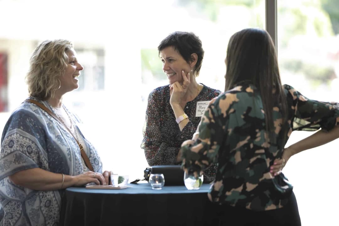 Three women are engaged in a conversation at a table with drinks in a bright setting. One woman, with curly hair and a patterned shawl, is smiling while leaning towards another woman wearing a floral blouse. The second woman, who has short, dark hair, is also smiling and appears to be listening attentively. The third woman, with long hair and a patterned top, is partially visible from the back, standing with her hands on her hips. A name tag is visible on the second woman, reading 'Mary Hew.'