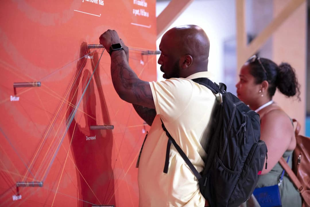 A man wearing a yellow shirt is interacting with a wall display that has an orange background and features three labels: 'Better,' 'Decreased,' and 'Worse.' He is placing a marker or similar item next to the 'Decreased' label, while a woman with a backpack stands in the background, observing the activity.
