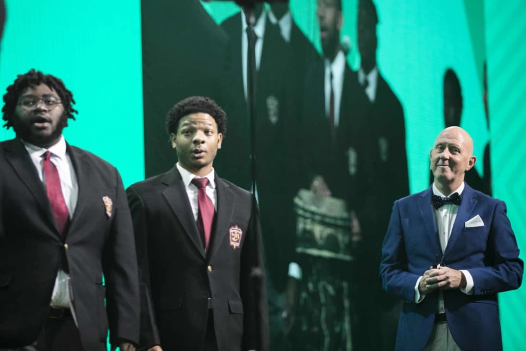 A performance scene featuring three individuals on stage, with two singers in dark suits and red ties, expressing emotion as they sing. In the foreground, a presenter in a blue suit and bow tie smiles while holding a microphone. Behind them, a large screen displays additional performers in the background against a green backdrop, adding to the festive atmosphere of the event.