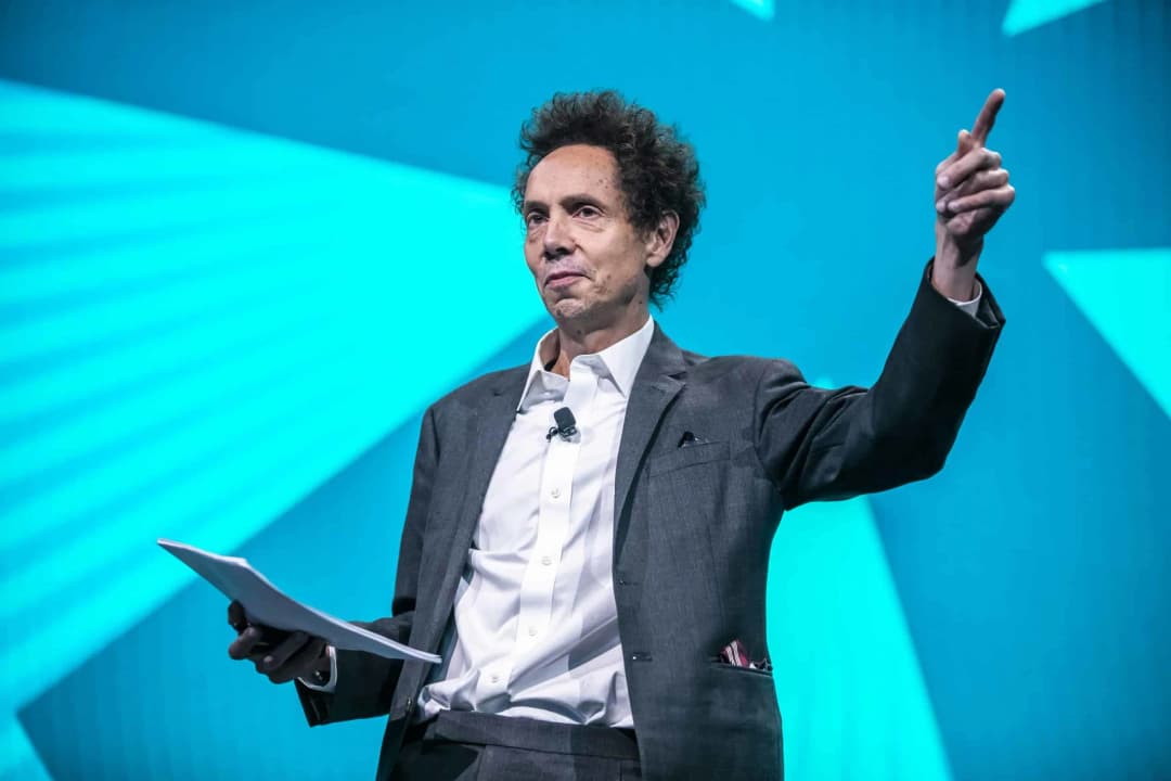 A speaker gesturing animatedly while holding a stack of papers, dressed in a gray suit and white shirt, stands in front of a bright blue background with geometric patterns. The scene captures a moment likely during a presentation or conference.