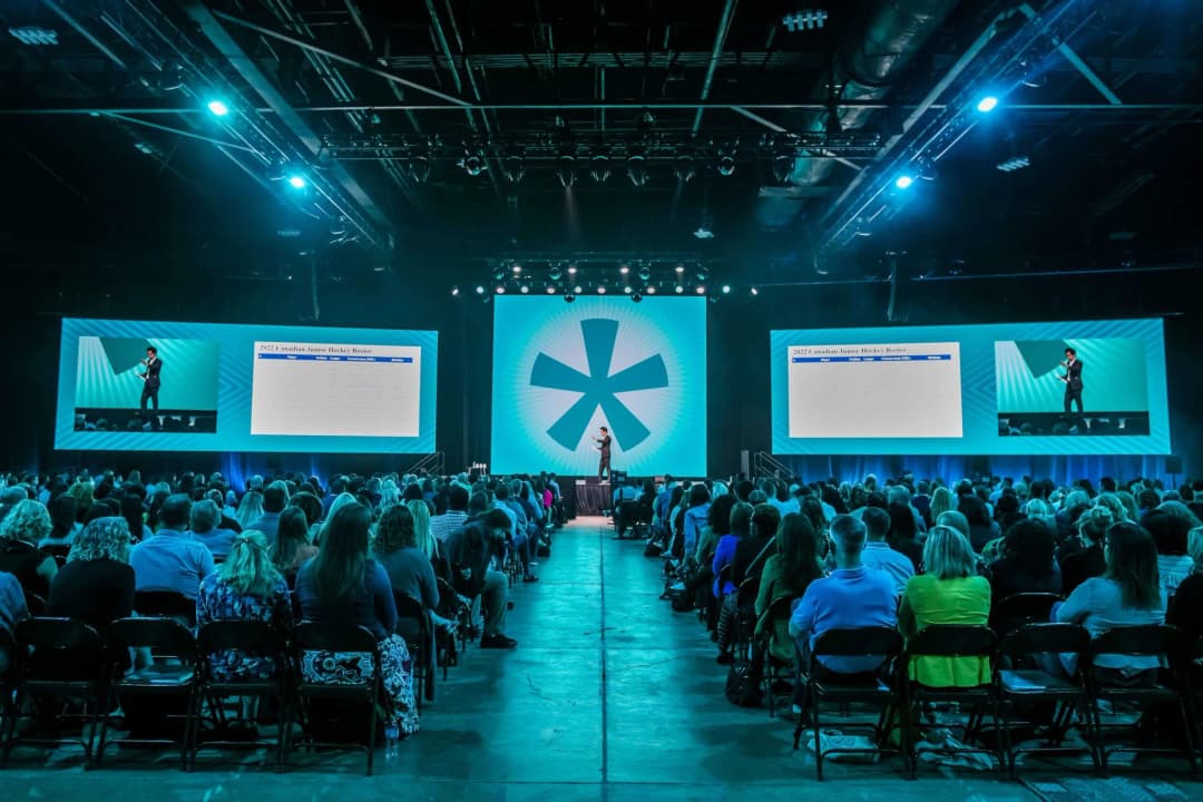 A speaker on stage presents to a large audience during a conference. Two large screens display the text '2022 Canadian Junior Hockey Roster.' The audience, seated in rows, appears engaged, with visible reactions to the presentation. The venue is well-lit, featuring a large symbol in the background, adding to the event's atmosphere.