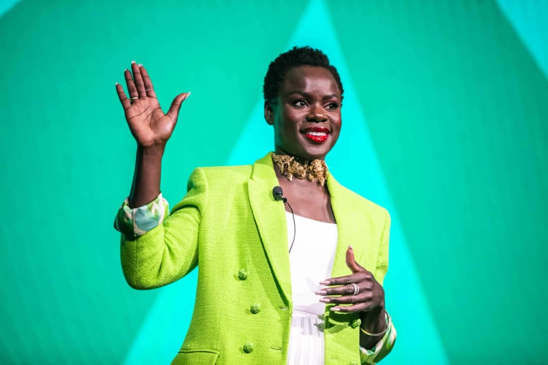 A woman stands on stage with her hand raised, smiling, wearing a vibrant green blazer over a white dress. She has a statement necklace and red lipstick, with a bright green background accentuating her presence.