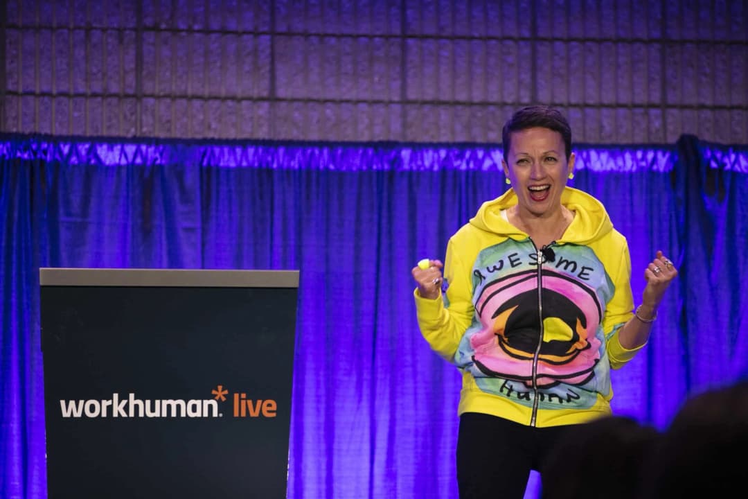 A woman enthusiastically speaks at the Workhuman Live event, wearing a colorful hoodie that has the word 'AWESOME' prominently displayed. She is gesturing expressively, conveying excitement. The backdrop features purple curtains, and there is a podium with the Workhuman Live logo visible.