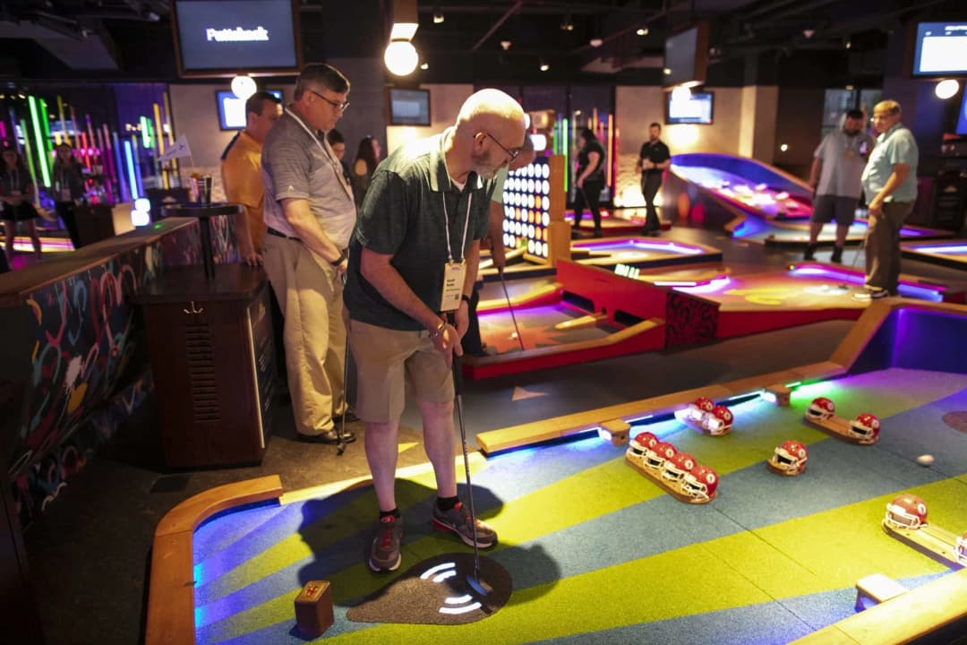 A group of people playing mini-golf at a vibrant indoor venue, featuring colorful lighting and various fun obstacles. One man, dressed in a dark shirt and shorts, is about to take a shot while two other participants observe. Behind them, a colorful bar area is visible, along with additional golfing setups in the background.