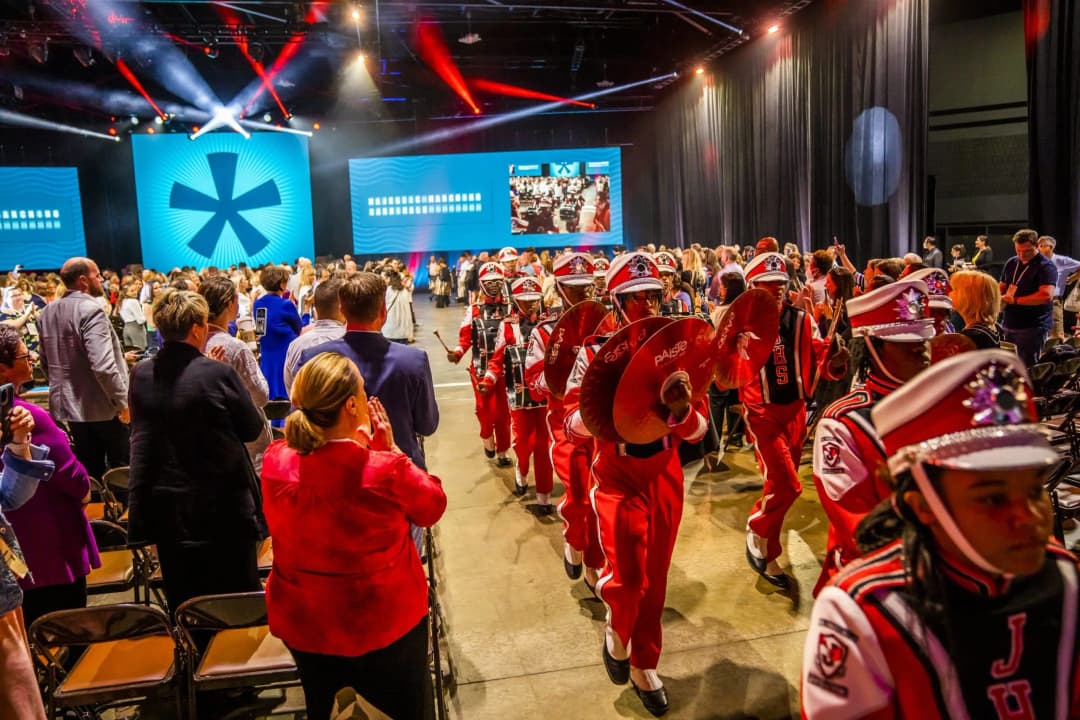A lively conference scene featuring an audience standing and applauding as a marching band in red uniforms parades through. The band members are playing cymbals and drums, creating an energetic atmosphere. In the background, large screens display visuals related to the event, enhancing the festive environment.