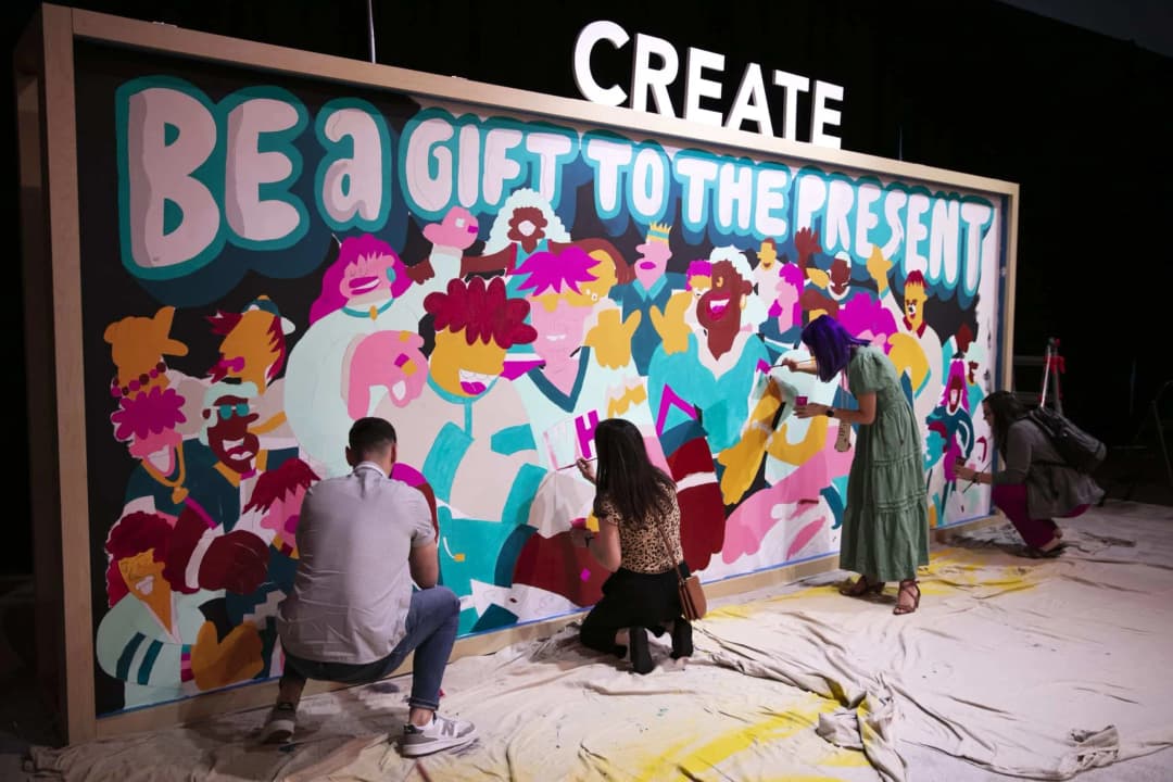 A vibrant mural titled 'BE A GIFT TO THE PRESENT' is being created by three individuals, each engaged in painting colorful figures and expressions. The scene features an assortment of lively characters with distinctive hairstyles, clothing, and expressions. Above the mural, the word 'CREATE' is prominently displayed. The artists are working on a large canvas positioned against a black backdrop, with a drop cloth covering the floor.