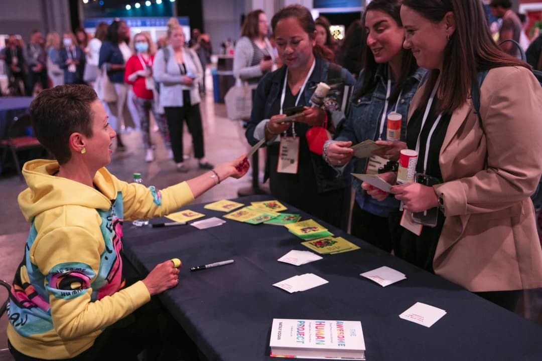 A woman in a colorful hoodie sits at a table, engaging with a group of attendees at an event. She is handing something to one of the attendees, while three others are looking at materials on the table, which features brightly colored cards and a book titled 'The Awesome Human Project' by Nataly Kogan. The background shows other attendees and a lively event atmosphere, highlighting interaction and networking.
