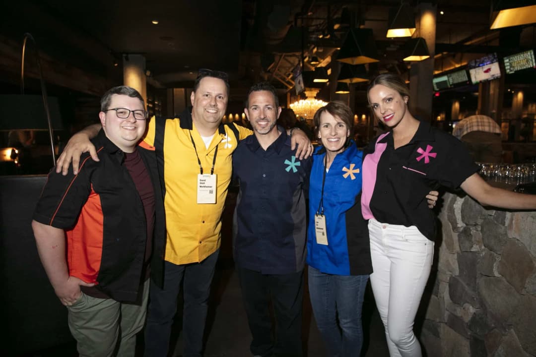 A group of five people is standing closely together, smiling and posing for the camera in a casual indoor setting. They are wearing brightly colored, matching shirts with different patterns: two men in black shirts with different color accents, one woman wears a blue shirt, another woman has a black shirt with pink accents, and one man has a yellow shirt. A name tag is visible on one of the men, identifying him as David. The background suggests a lively atmosphere with soft lighting and hints of decor typical of a social gathering or event.