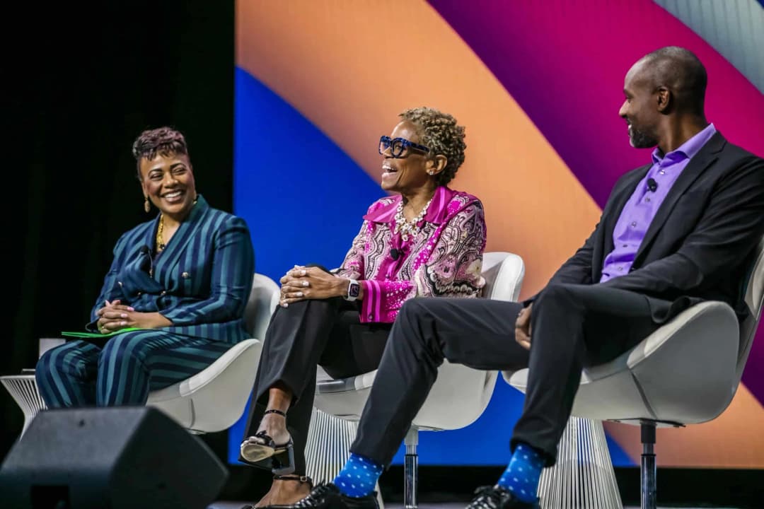 A panel of three speakers at a Workhuman event, engaged in conversation and smiling. The first speaker, a woman in a striped suit, is sitting on the left, while the second speaker, a woman wearing a colorful blouse, sits in the middle. The third speaker, a man in a black suit with distinctive blue socks, is on the right. They are seated in modern chairs, positioned against a backdrop of vibrant colors.