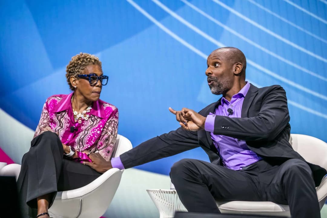 A man in a black suit and purple shirt is animatedly speaking while gesturing with his hand. Next to him, a woman with short, curly hair wearing large glasses and a patterned pink blouse listens attentively. They appear to be engaged in a discussion on stage, set against a blue background with light patterns.