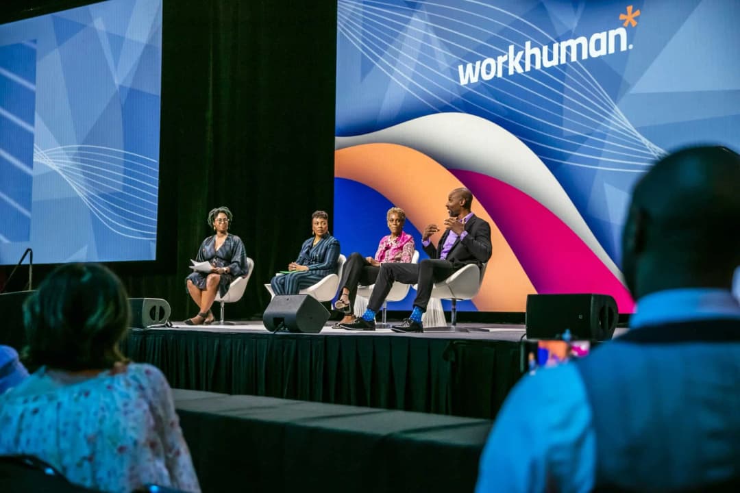 A panel of four speakers is seated on stage at a Workhuman event, with one speaker actively gesturing while speaking. The backdrop features the Workhuman logo and a colorful design. Attendees in the foreground are facing the stage, and one individual is holding a camera, capturing the moment.