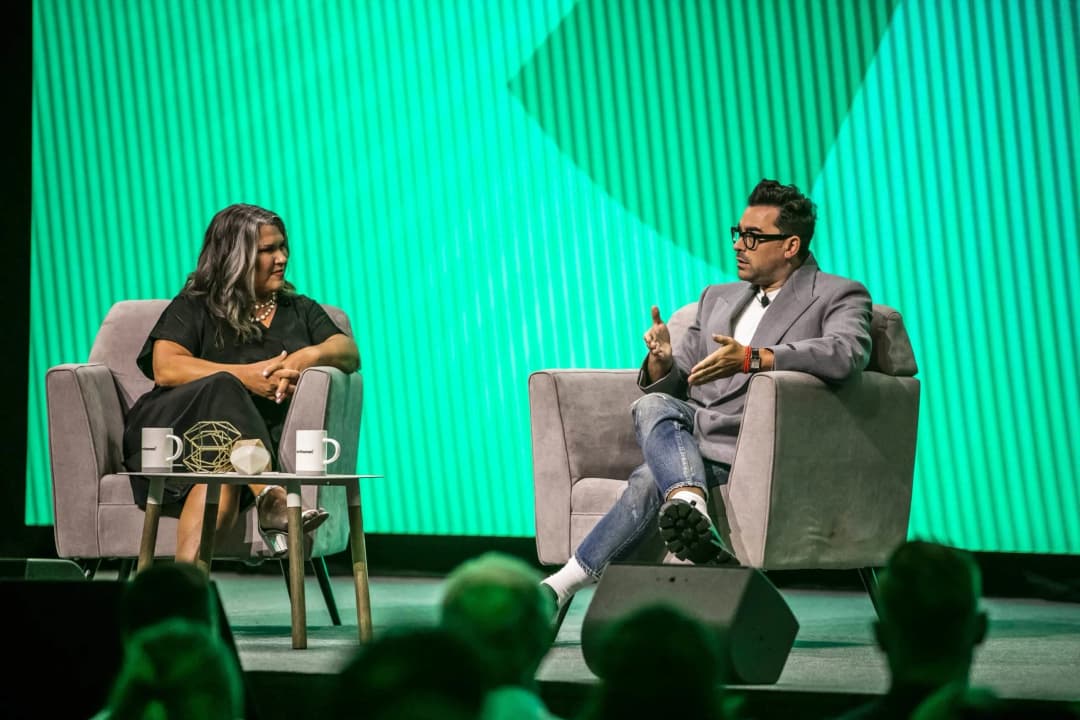 A discussion panel featuring two speakers seated in gray armchairs on stage. The speaker on the left, a woman with long, black hair, is wearing a black dress. The speaker on the right, a man with short hair and glasses, is dressed in a gray blazer, white t-shirt, and jeans. A small table in front of them holds two coffee cups and a decorative object. The background is a vibrant green with vertical stripes, and part of the audience is visible in the foreground, focusing on the speakers.