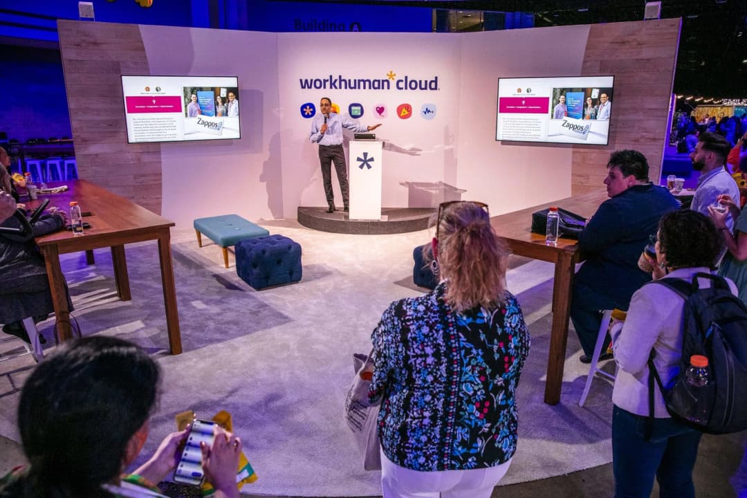 A speaker presenting at a Workhuman event in front of a display featuring the Workhuman cloud logo and the Zappos brand. Attendees are seated at a wooden table, with a blue ottoman nearby. The atmosphere is lively, with a colorful backdrop and screens showing information.