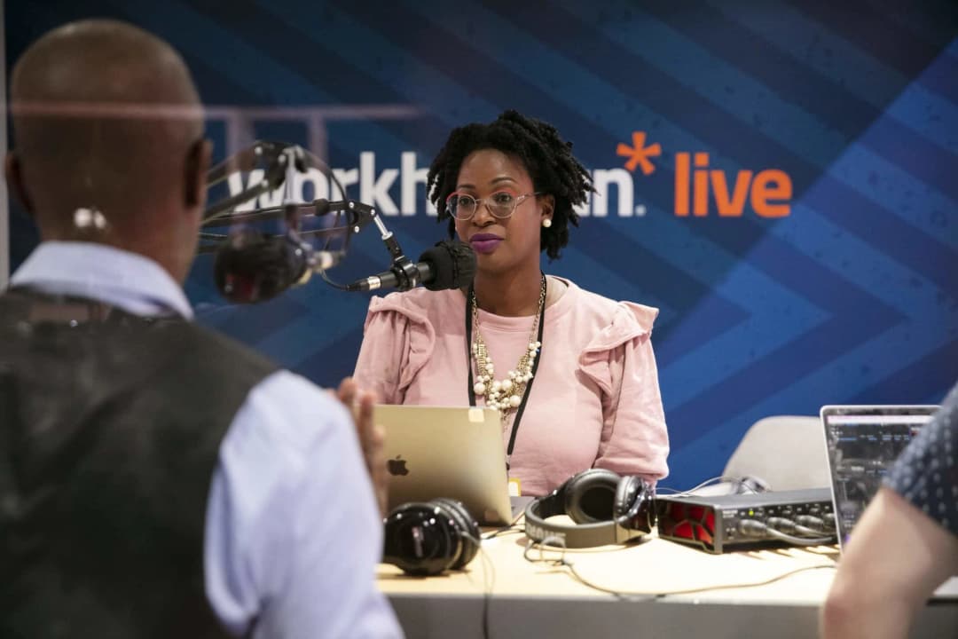 A woman with curly hair, wearing glasses and a pink top, is sitting at a table in a podcast studio, speaking into a microphone. She is surrounded by audio equipment and a laptop. In the background, there is a blue backdrop with the Workhuman logo and the word 'live' prominently displayed. A man, seen from behind, is listening to her, creating an engaging atmosphere.