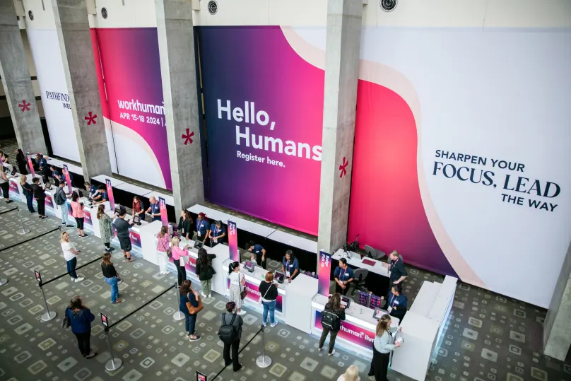A view of the registration area for the Workhuman event, featuring large banners that say 'Hello, Humans' and 'Sharpen Your FOCUS, LEAD THE WAY,' alongside the dates 'APR 15-18, 2024.' Attendees are standing in line at the registration desks while others are engaged with the staff members to their right.