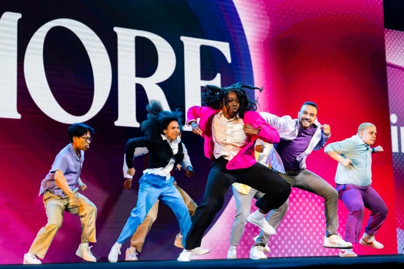 A group of dancers performing on stage, showcasing dynamic movements and expressions. They are dressed in vibrant clothing, with one dancer in a bright pink jacket leading the performance. The backdrop features bold text that reads 'MORE,' setting a lively atmosphere.