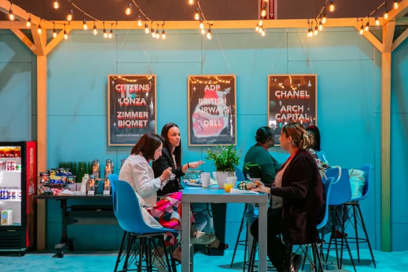 A group of four women and one man are seated around a table in a colorful, well-lit space adorned with string lights. They are engaged in conversation, with some enjoying food and drink. Behind them are several framed posters featuring company names like 'ADP,' 'British Airways,' 'Chanel,' 'Arch Capital,' and others. The backdrop is painted a vibrant blue, creating a cheerful atmosphere.