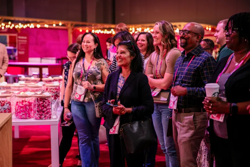 A group of happy attendees, mostly women, are gathered at a lively event. They are standing close together, smiling and interacting with each other. In the background, there are tables filled with decorative boxes and soft lighting, creating a warm atmosphere. Some participants are wearing lanyards with name tags, while one person holds a coffee cup.