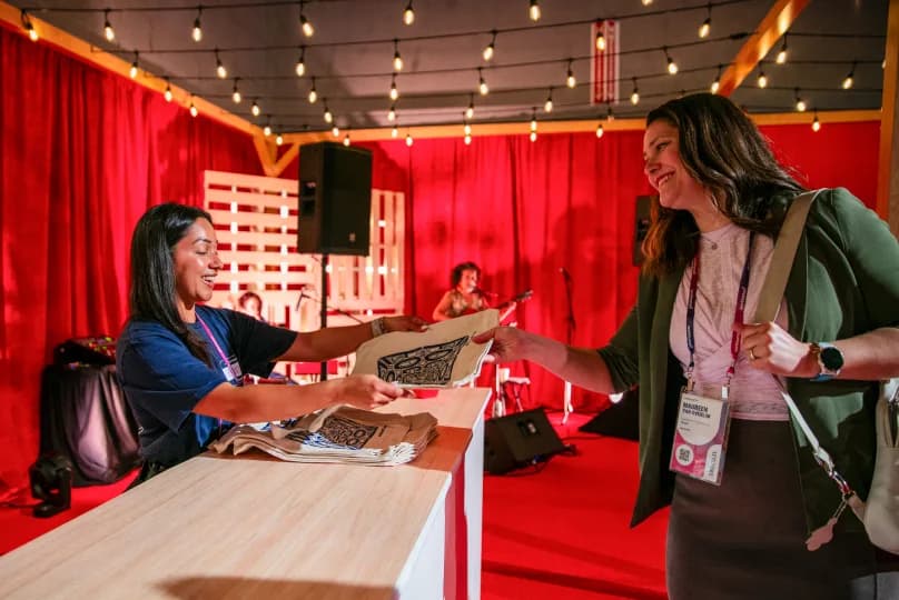A woman in a blue t-shirt hands a wrapped gift or item to another woman dressed in a green jacket and gray skirt, who is smiling and holding a bag. The background features red curtains, string lights, and a small live music setup with musicians. The scene suggests a warm, festive atmosphere, likely at an event.