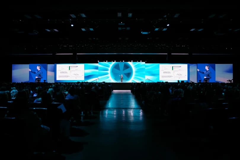 A large audience is seated in a conference hall, facing a speaker on stage. The speaker is gesturing towards a large projection screen that displays colorful graphics and data. The venue is brightly lit, and various visual elements enhance the presentation atmosphere.