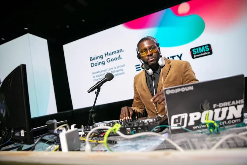 A DJ wearing headphones and sunglasses stands behind a mixing console at the Workhuman Live event. A large screen behind him displays the phrases 'Being Human. Doing Good.' and mentions charitable support linked to Workhuman. The overall atmosphere features a vibrant setup, including colorful graphics on the backdrop.