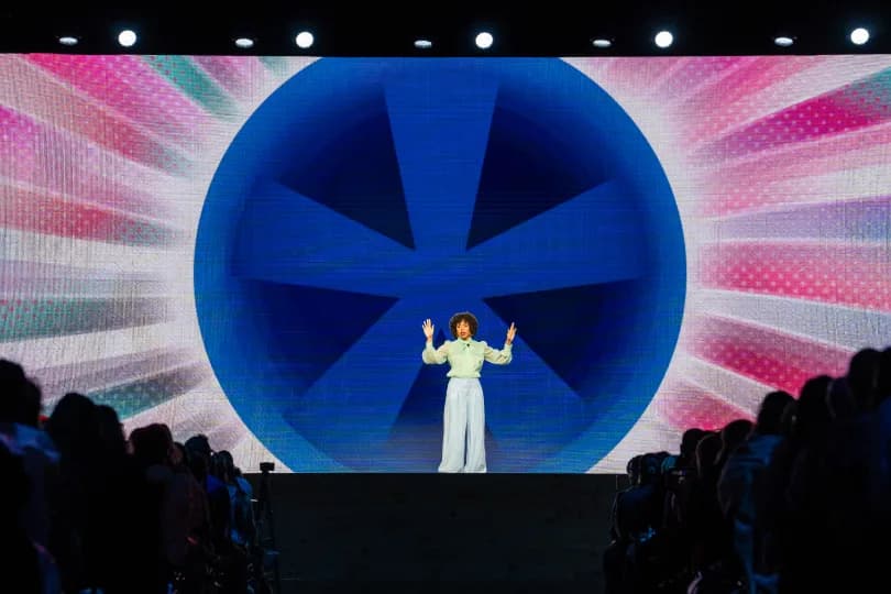 A performer stands on stage with arms raised, presenting to the audience. Behind them is a vibrant backdrop featuring a large blue circle with radiating colors and patterns. The audience, not fully visible, appears engaged in the performance.